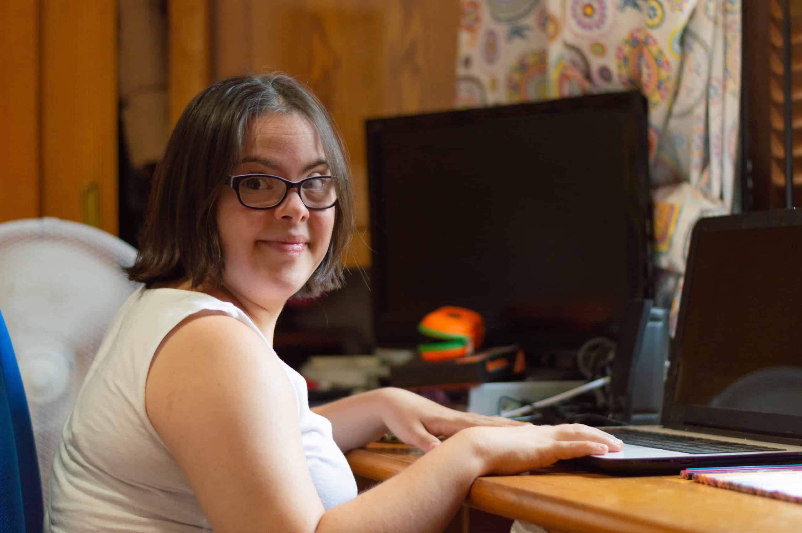 Young adult female with Down syndrome sitting in the bedroom of her home, using a laptop computer and smiling at the camera.