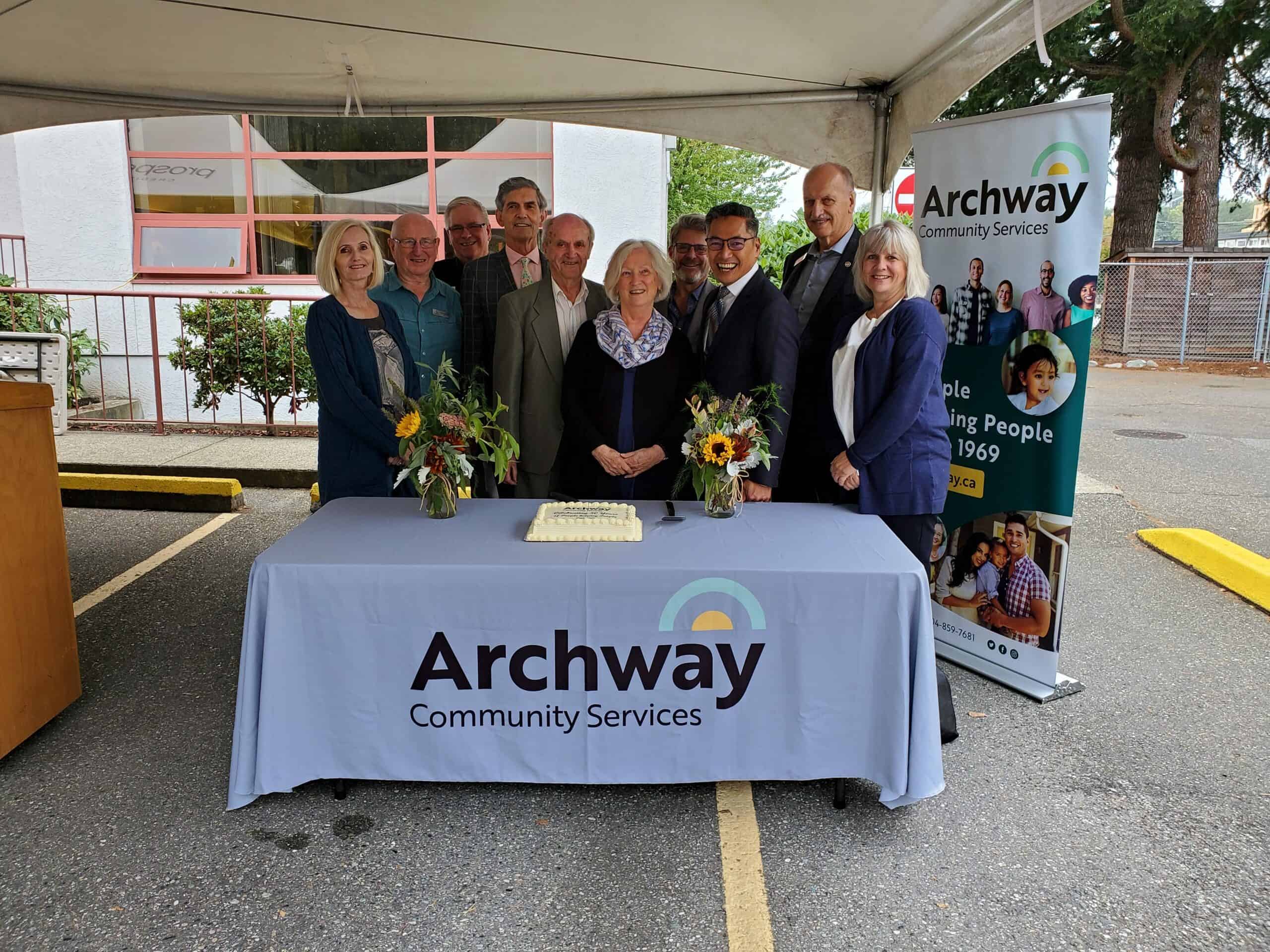 Archway staff stand behind a table branded with the Archway logo, with cake and flowers upon it, outside under a large canopy.