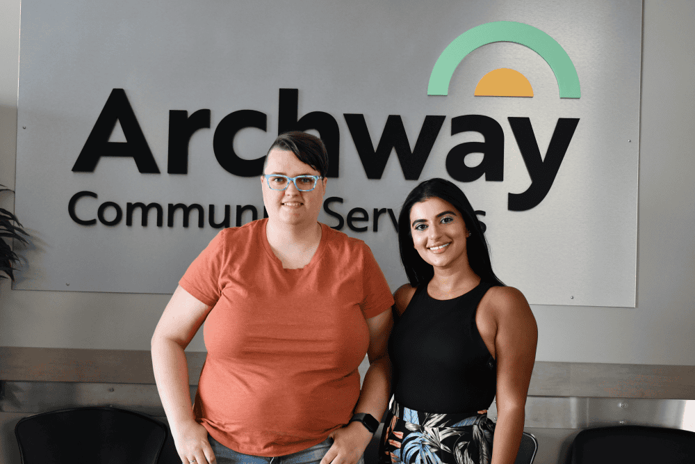 Two young adult females pose in front of the Archway Community Services wall logo