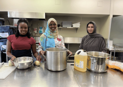 three young adult women smile in industrial kitchen with large cooking pots on table in front of them