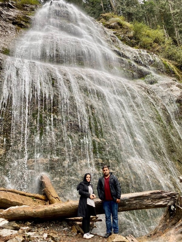 Male and female adult stand in front of a waterfall looking at the camera