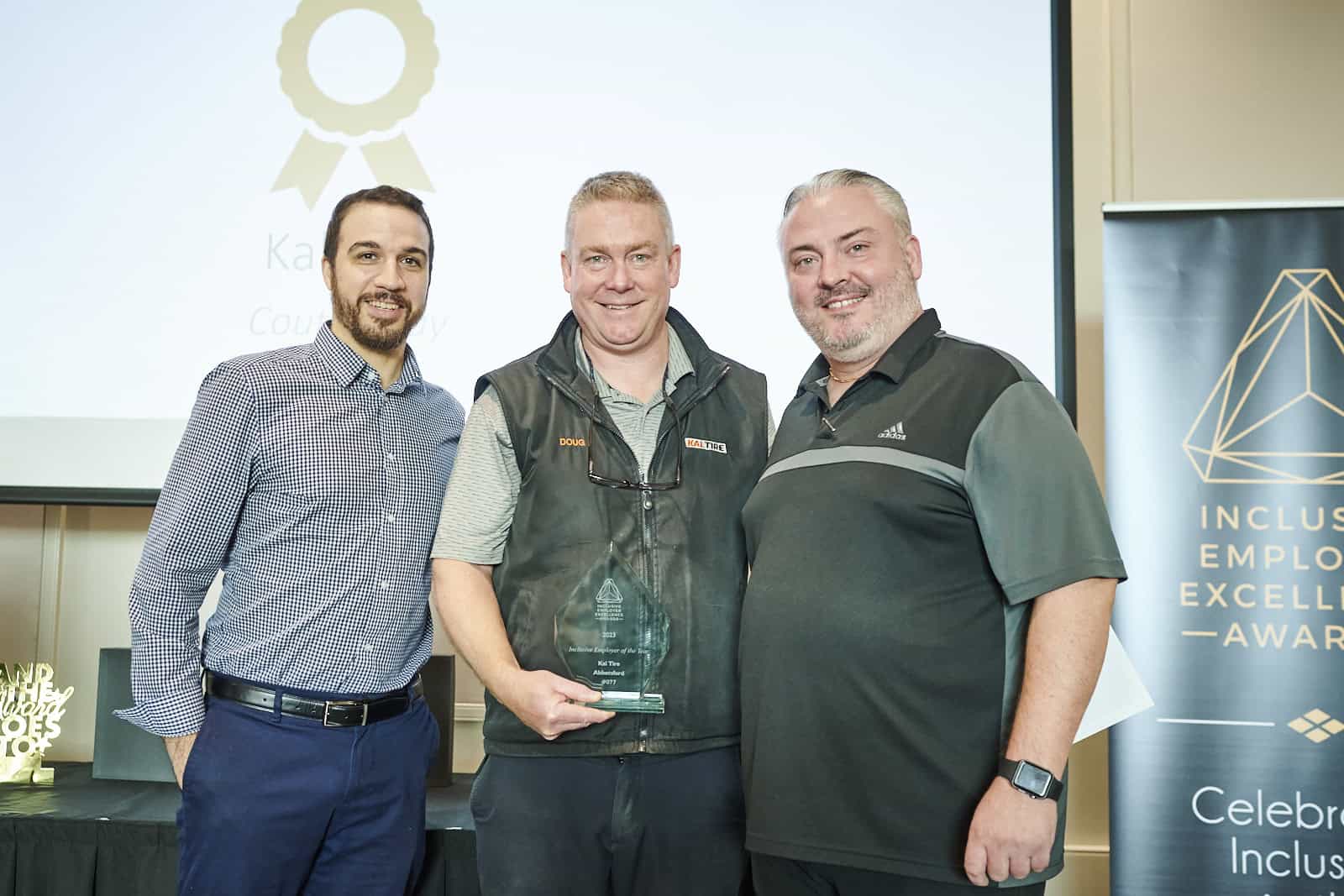 three men on smiling at camera standing next to each other. The man in the middle holds a glass trophy.