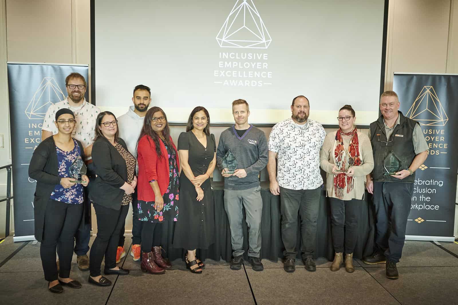 Group of people on stage smiling at the camera with the inclusive employer excellence logo behind them on a projector screen