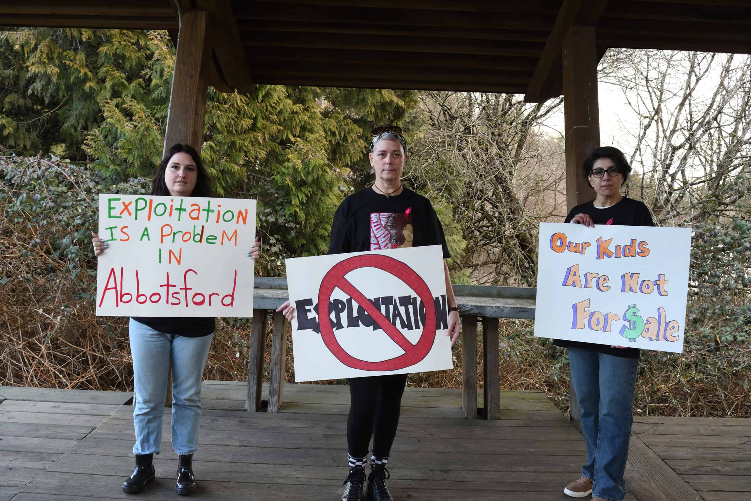 Three people stand abreast holding one poster each that read "Exploitation is a problem in Abbotsford", "Our kids are not for sale" and a large prohibited symbol on top of the word "Exploitation'