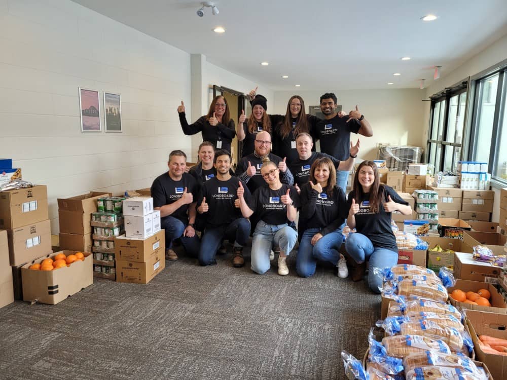 Group of volunteers in a room with food and boxes smiling at the camera wearing dark blue shirts