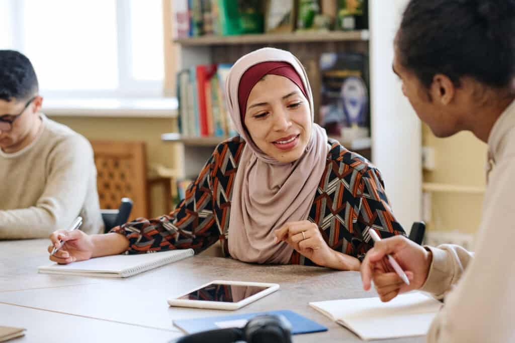 woman wearing hijab sitting at table in library taking look into notebook of her classmate during lesson