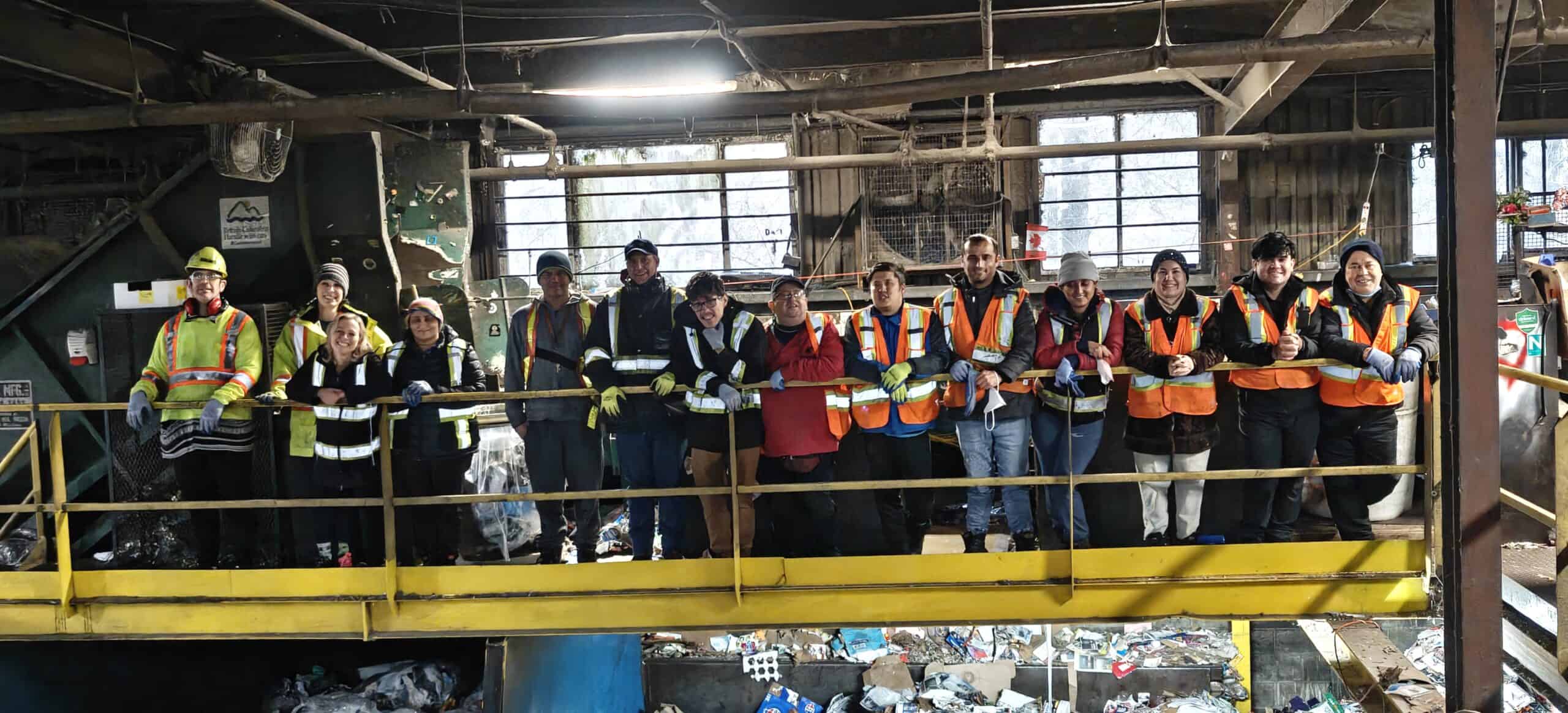 Group of people stand abreast behind a railing in a recycling facility